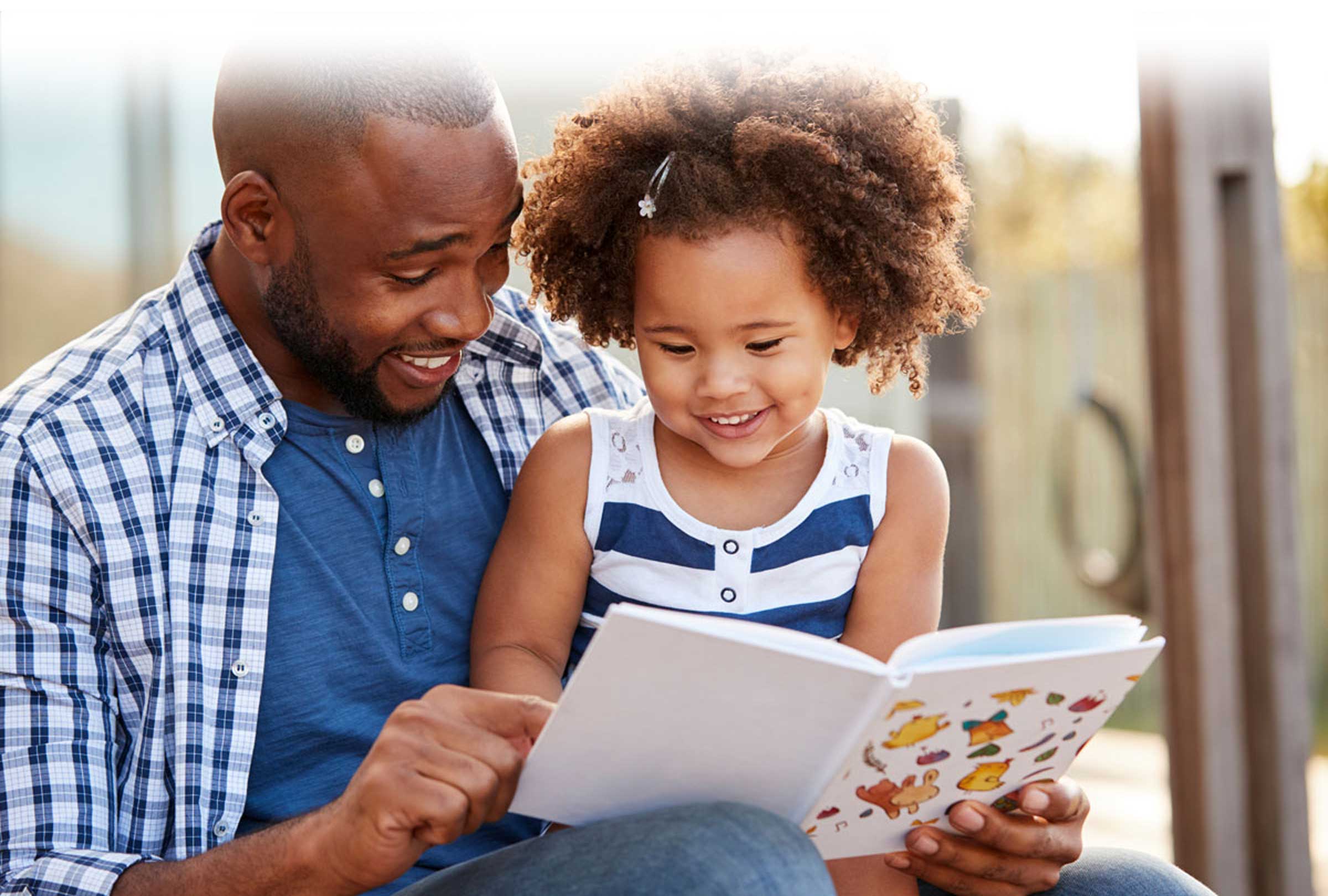 Dad with daughter reading book, smiling, outdoors